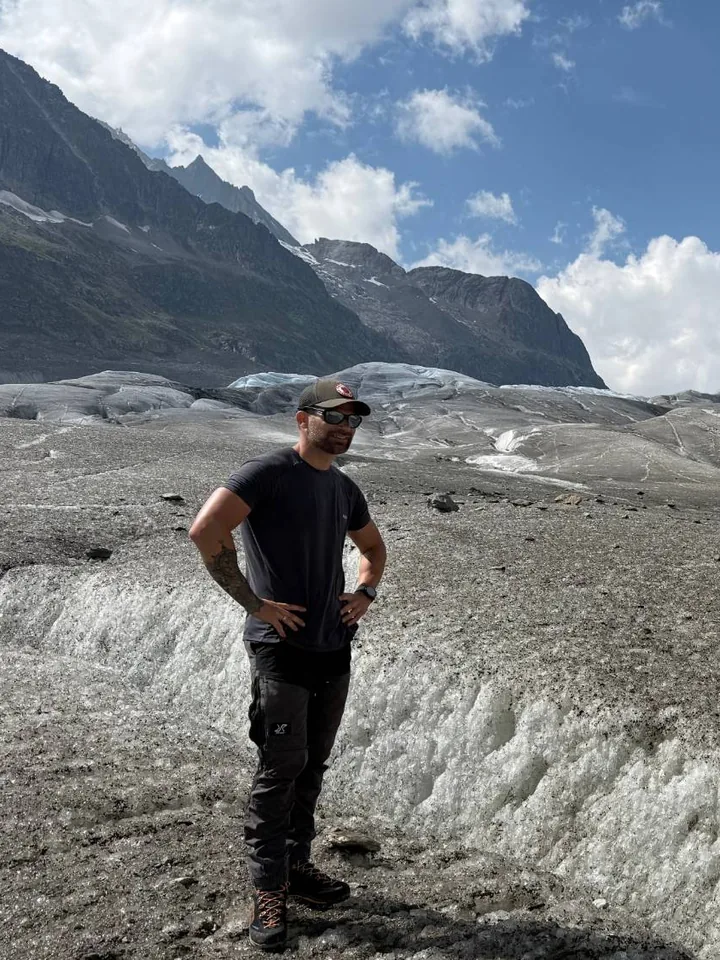 Youri standing on a debris-covered glacier, with mountains and clouds behind him.